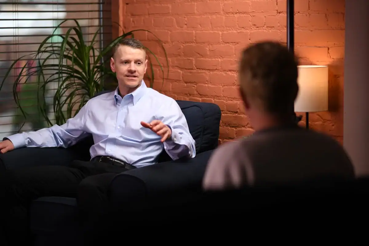 A man in a light blue shirt sits on a dark sofa, gesturing while discussing OCD treatment in Ramsey County, MN with another person in a warmly lit room featuring a brick wall, lamp, and leafy plant.