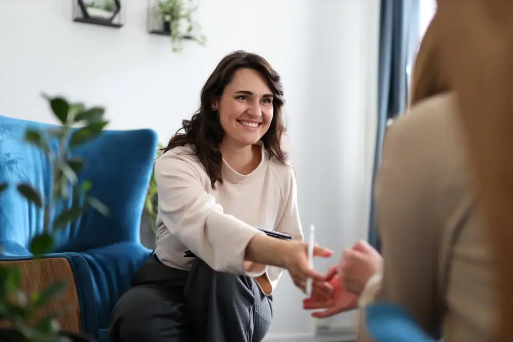 A smiling woman sits on a blue chair, leaning forward to greet someone in a cozy, well-lit room with plants—an inviting scene that could represent the warm environment of OCD Therapy Texas and Minnesota.