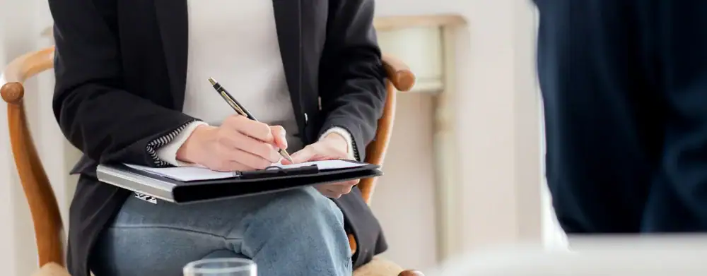 A person in a suit jacket and jeans sits on a chair, holding a clipboard and writing with a pen, possibly during an ERP Therapy Texas and Minnesota session or counseling; another blurred figure is visible in the foreground.