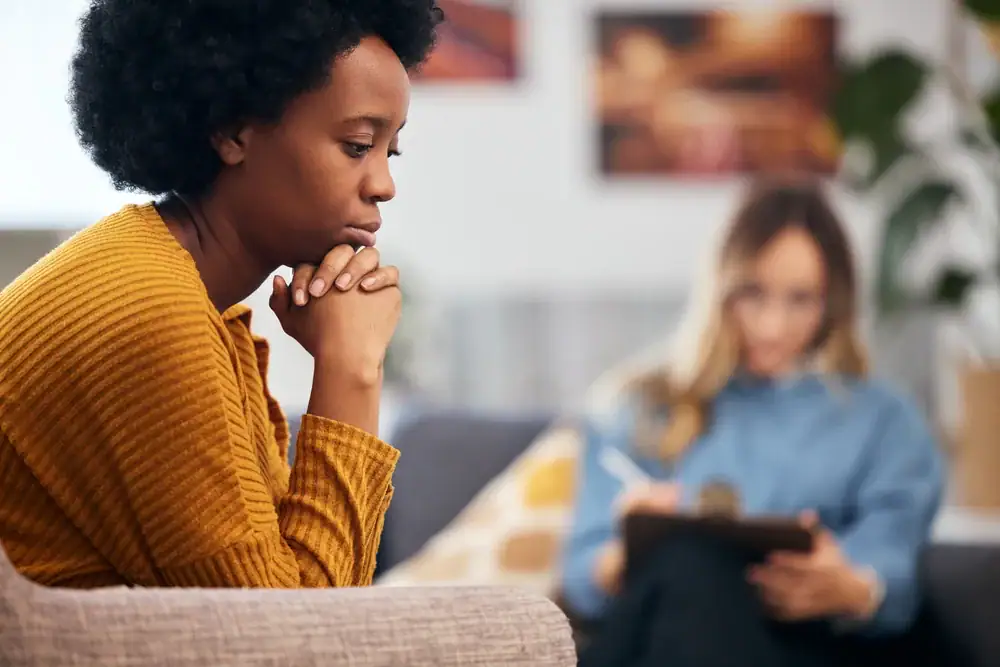 A woman in a yellow sweater sits with clasped hands, looking thoughtful or concerned, while another person in the background takes notes—suggesting an OCD Therapy Texas and Minnesota session focused on care and understanding.
