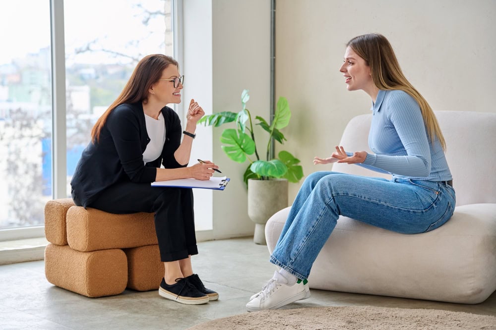 A woman with a clipboard and pen sits on a stool, attentively listening to another woman in a blue sweater and jeans discussing her experience with OCD Therapy Texas and Minnesota in a bright, modern room filled with plants.