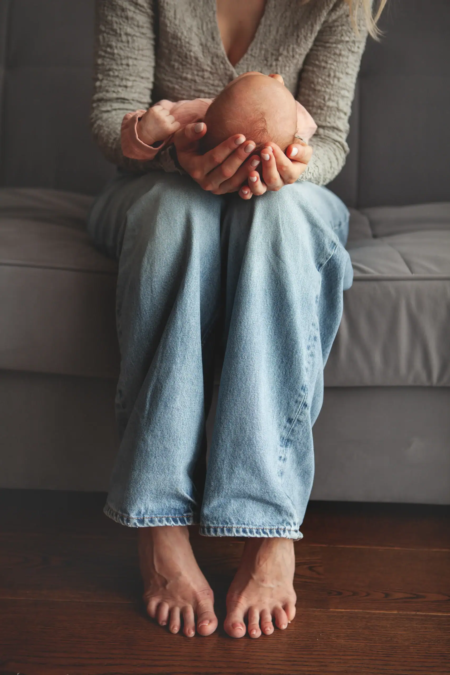A person sits barefoot on a gray couch, wearing loose blue jeans and a sweater, with their hands clasped around their knees. Their face is not visible, reflecting the quiet moments often found during OCD therapy Texas and Minnesota sessions.