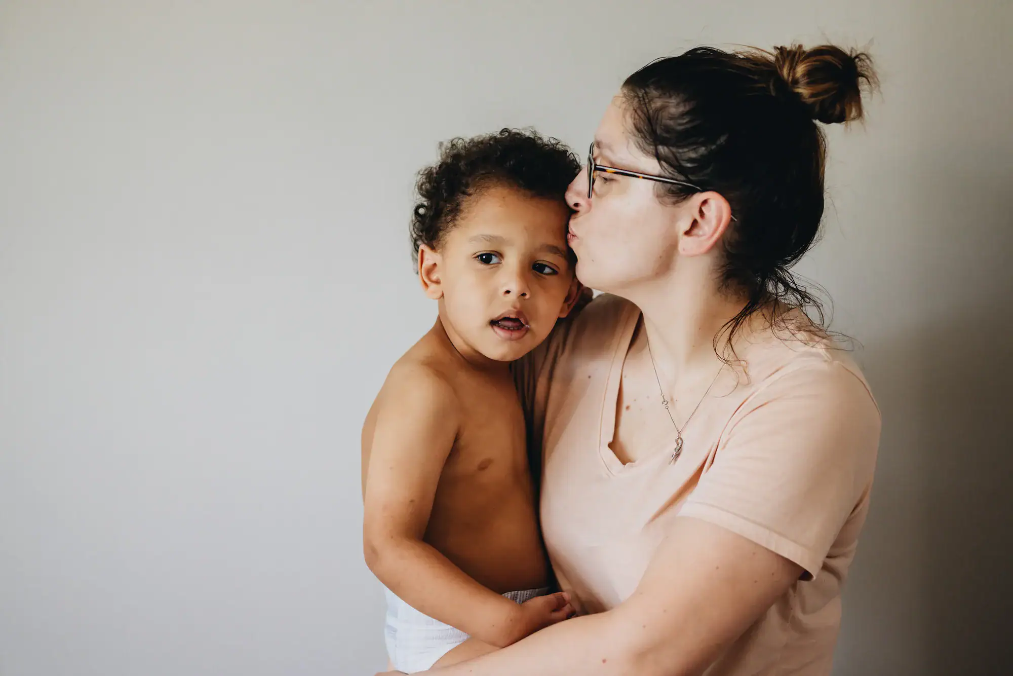 A woman in glasses and a light pink shirt kisses a young child on the head. The child, shirtless and in a diaper, looks at the camera while being held close—capturing warmth that reflects the care of OCD therapy Texas and Minnesota offer.