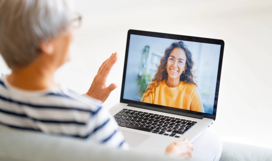 An older woman with short gray hair waves at a young woman on a laptop screen during a video call. The young woman wears glasses and a yellow sweater, smiling brightly.